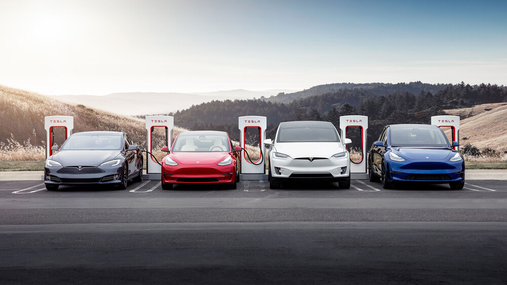 Die vier Tesla Modelle Model 3 Model S Model X Model Y stehen auf einem Parkplatz mit Supercharger Ladesäulen vor schöner Landschaft