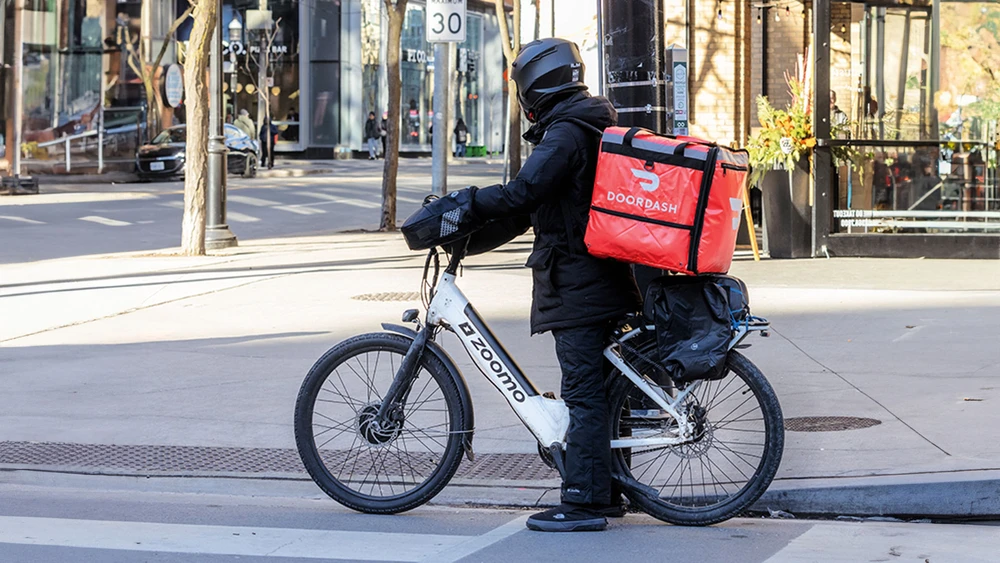 Ein DoorDash-Essenslieferkurier auf einem Elektrofahrrad oder E-Bike mit dem markanten roten Rucksack auf einer Straße in der Innenstadt von Toronto