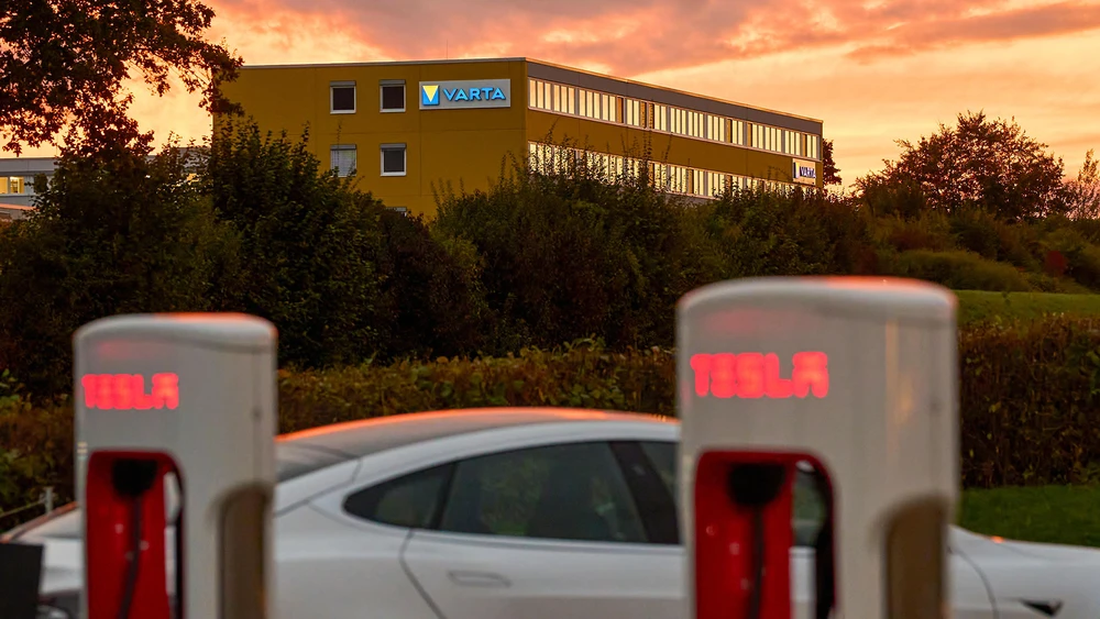 Tesla Ladesäulen vor Gebäude mit beleuchtetem Varta Logo vor rosa roten Wolken